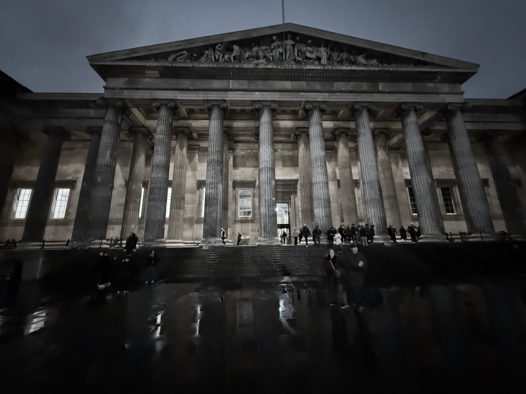 A black and white photo of the British Museum at early evening. The night was rainy. The shot is at a Dutch angle as the museum entrance towers above