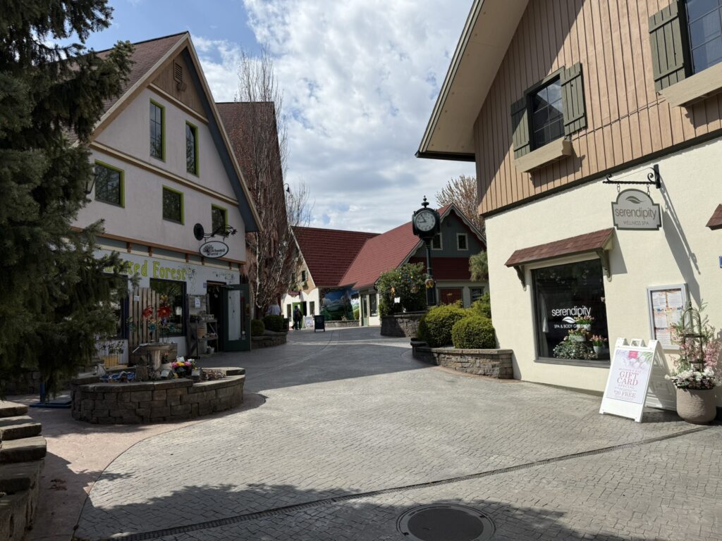 Photo of a winding street, looking uphill. There's water fountain in the foreground at the left part of the photo. There are three people in the far distance. The scene is sunny, nearly mid-day. On each side are three-story buildings meant to look like German houses, each with a sign for the store within hanging just above the first story line. Everything in Frankenmuth is built for tourism and isn't genuine, so everything has a slightly off feeling. It looks a lot like the fake heaven created in the TV show "The Good Place"